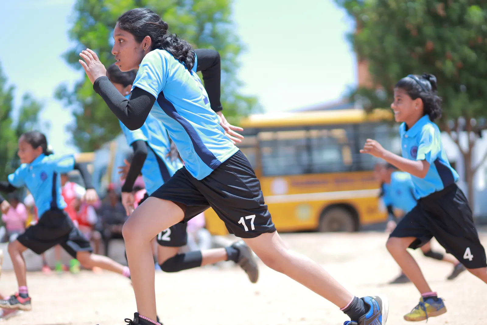 Girls sprint across the sports field, a school bus behind them